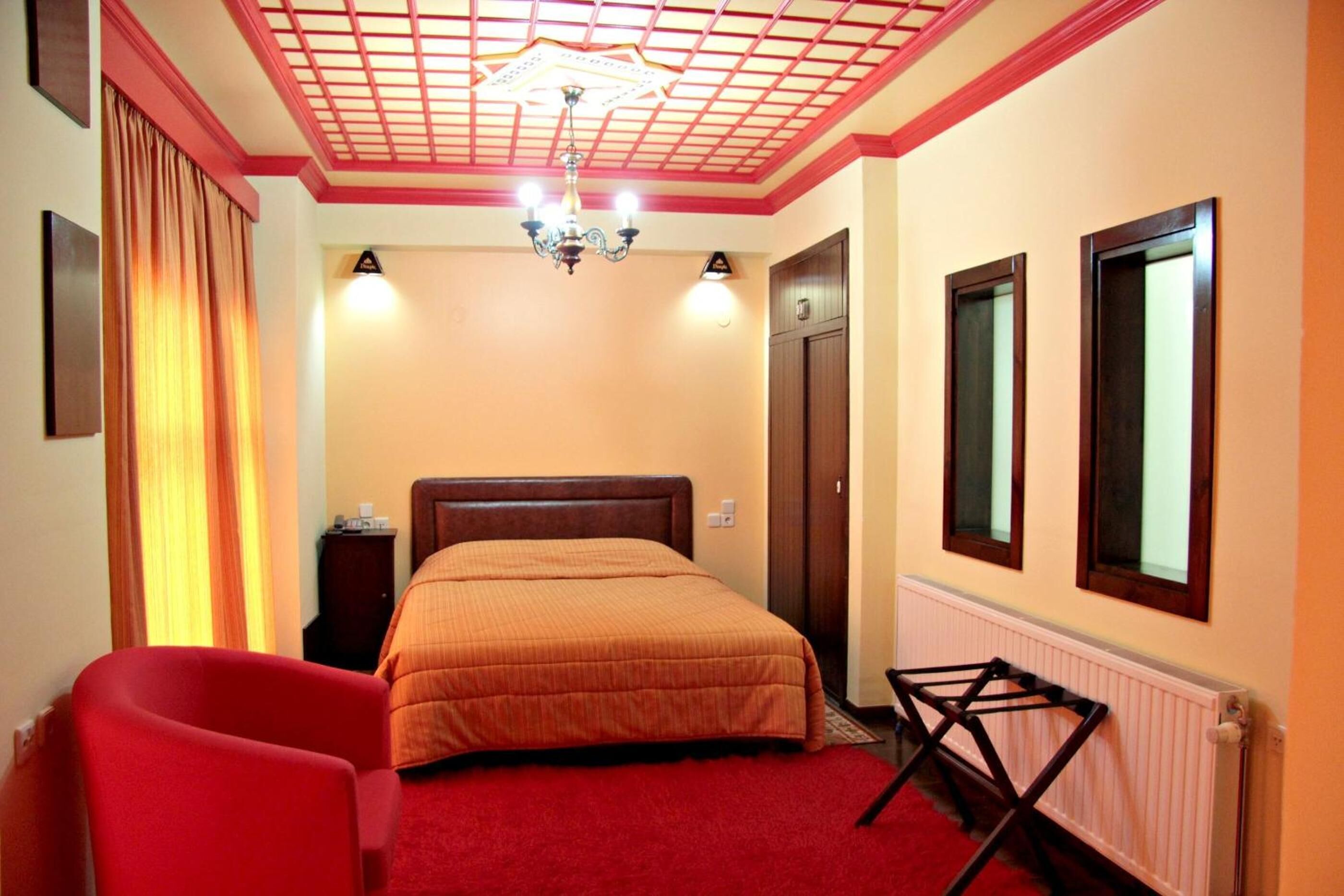 Een hotelkamer met een bed, een rode stoel, een houten nachtkastje, een kroonluchter en een radiator.