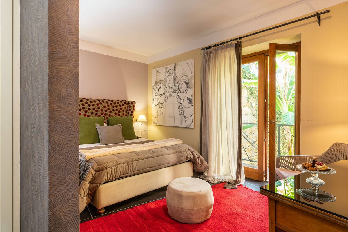 Une chambre à coucher avec un lit, un tapis rouge, un pouf rond, une table à manger avec une lampe et une fenêtre avec des rideaux.