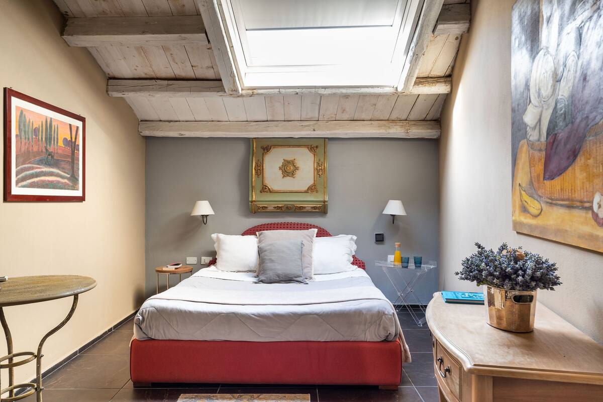 Une chambre avec un puits de lumière, un lit avec une literie rouge et blanche, une petite table et une table de chevet avec une lampe.
