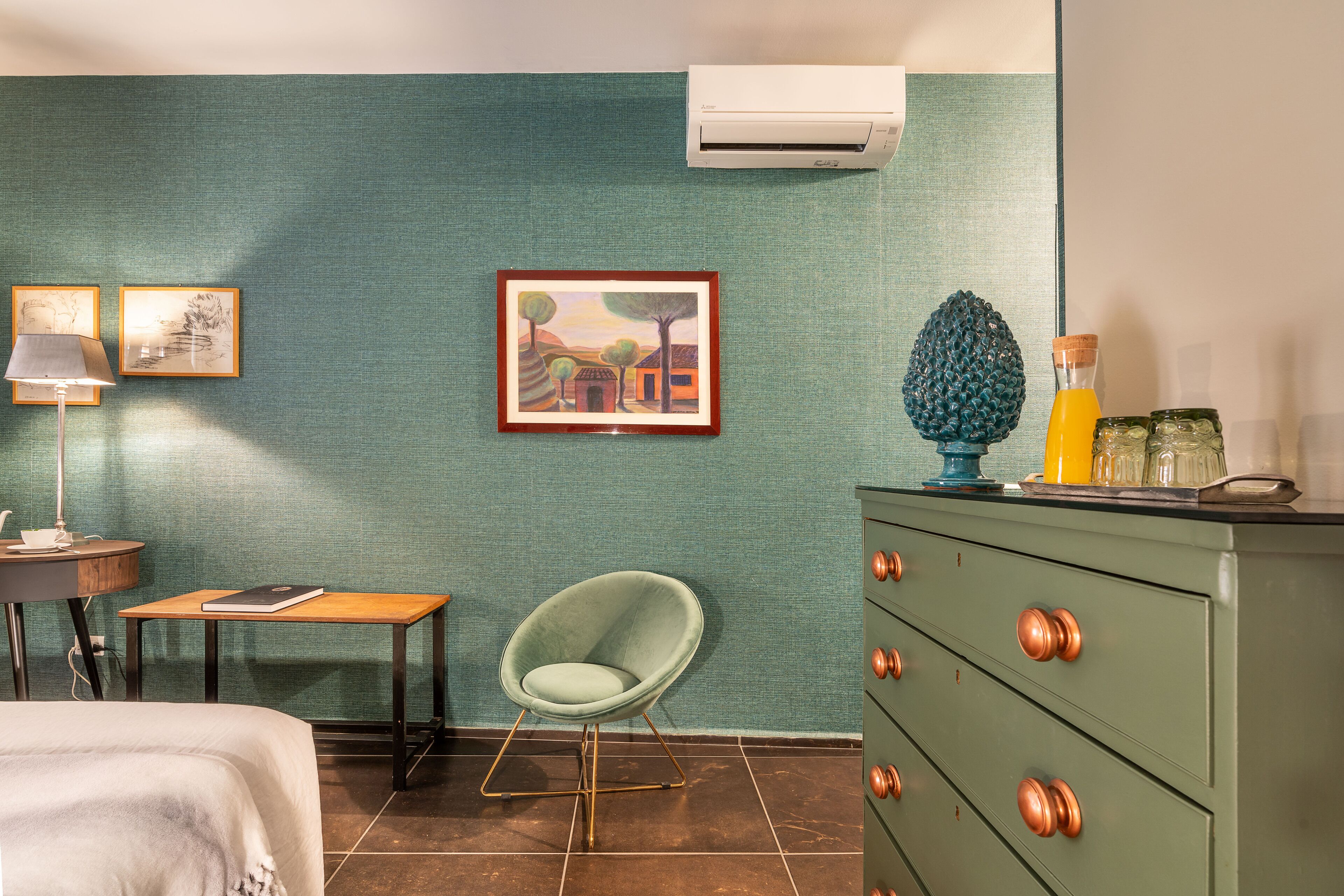 Habitación de hotel con paredes verde azuladas, una silla verde, una mesa de madera con una libreta, un aparador verde con pomos de cobre y un cuadro enmarcado.