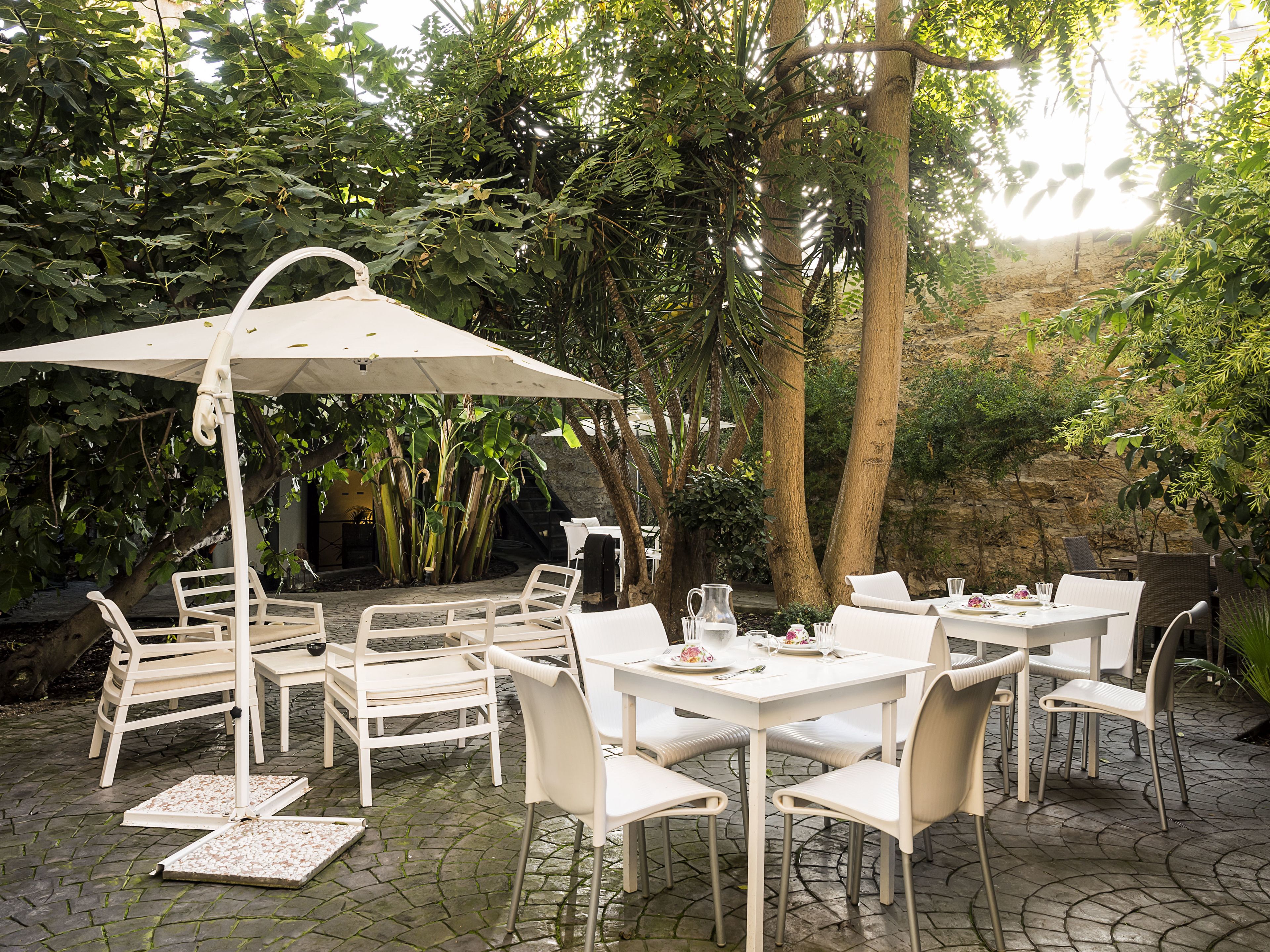An outdoor patio with white tables and chairs, surrounded by lush greenery and trees.