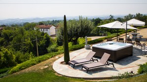 A hot tub with two lounge chairs, surrounded by a stone patio, with a view of hills and a house in the background. - Cascina Bellavista (Tigliole)