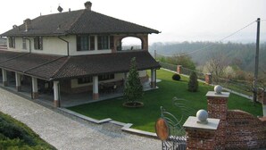 A large two-story house with a covered patio, a well-maintained lawn, and a brick wall with decorative elements. - Cascina Bellavista (Tigliole)