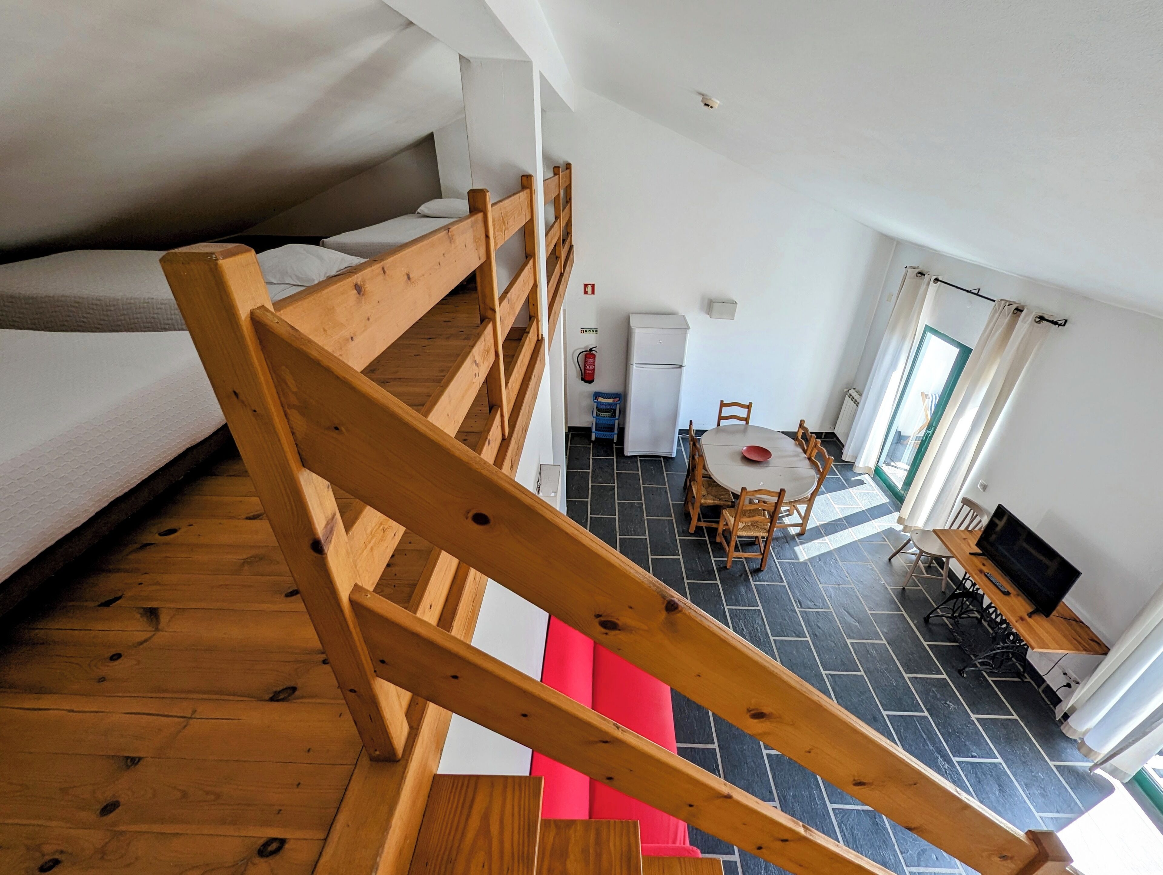 Un dormitorio tipo loft con escalera de madera, una kitchenette pequeña, un rincón comedor con mesa y sillas, y un televisor.