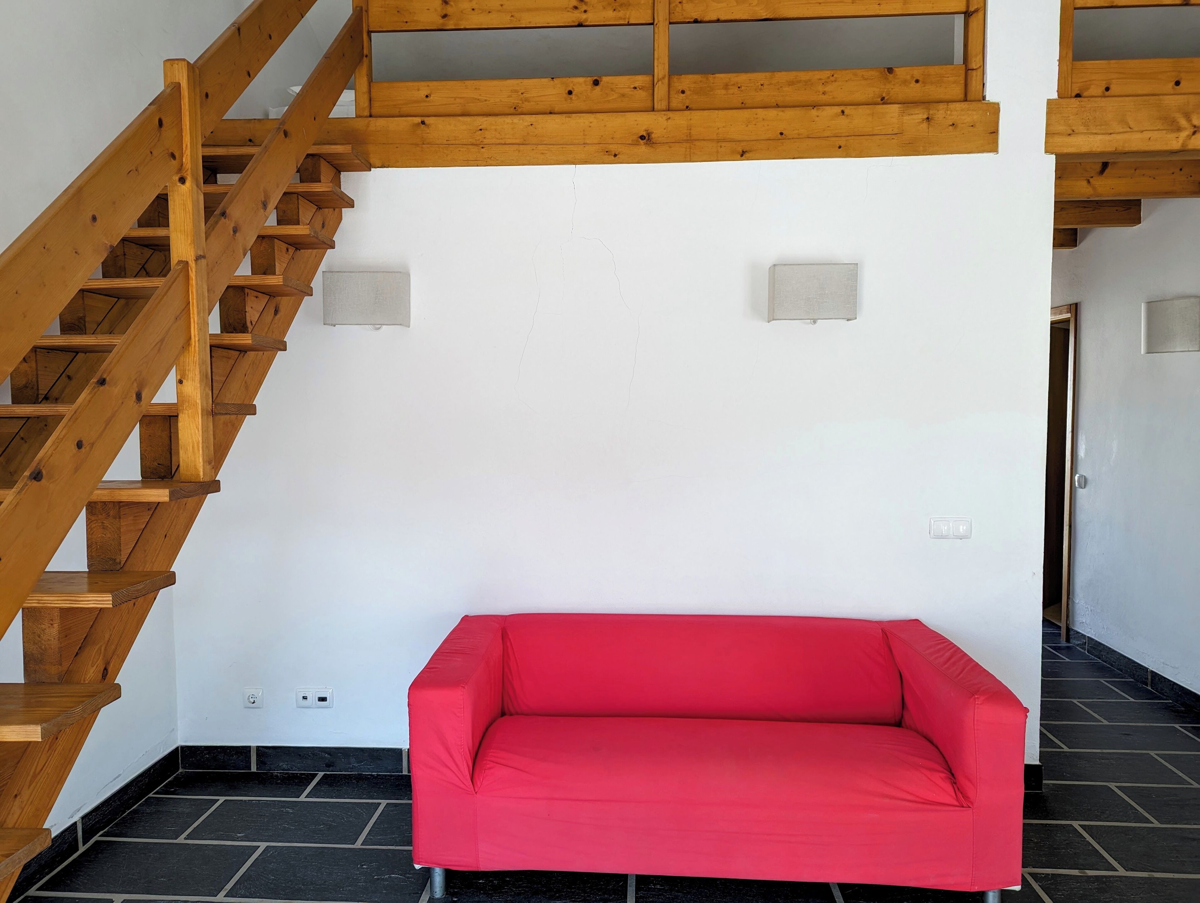 Une pièce avec un escalier en bois, un canapé rouge et un mur blanc orné de deux interrupteurs.