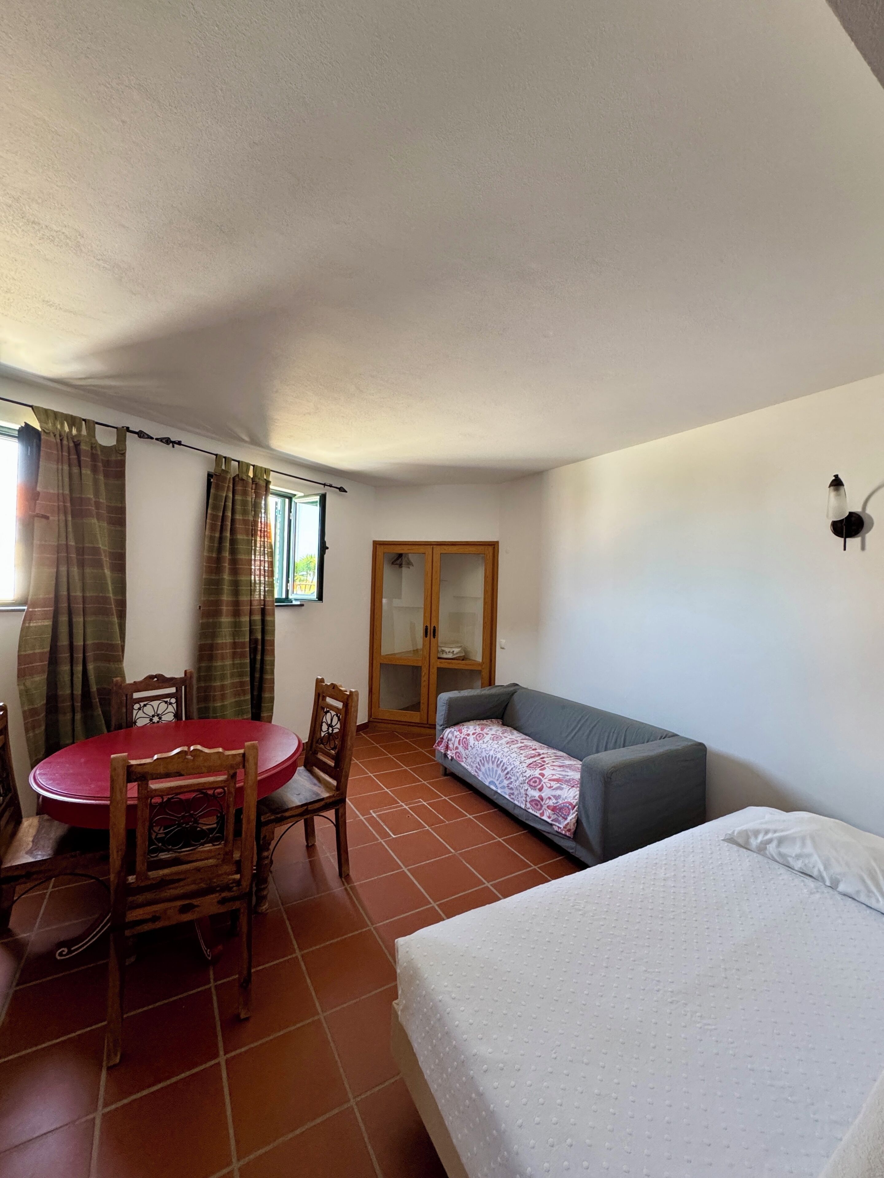 A room with a bed, a sofa, a dining table with chairs, a wooden cabinet, and a window with curtains.
