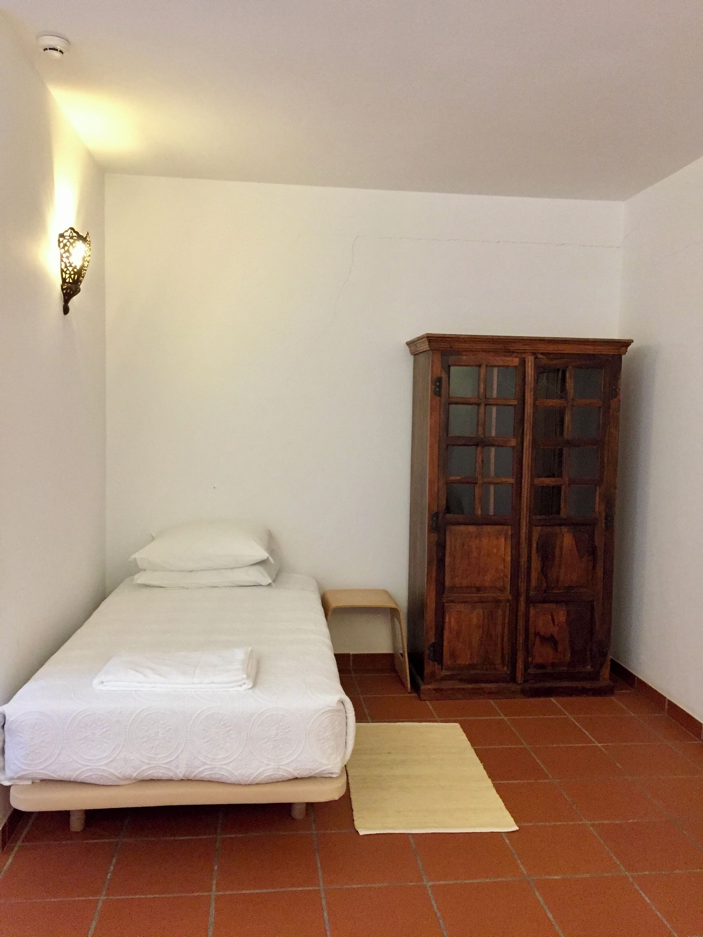 A small, clean room with a single bed, a wooden wardrobe, and a wall-mounted light fixture.