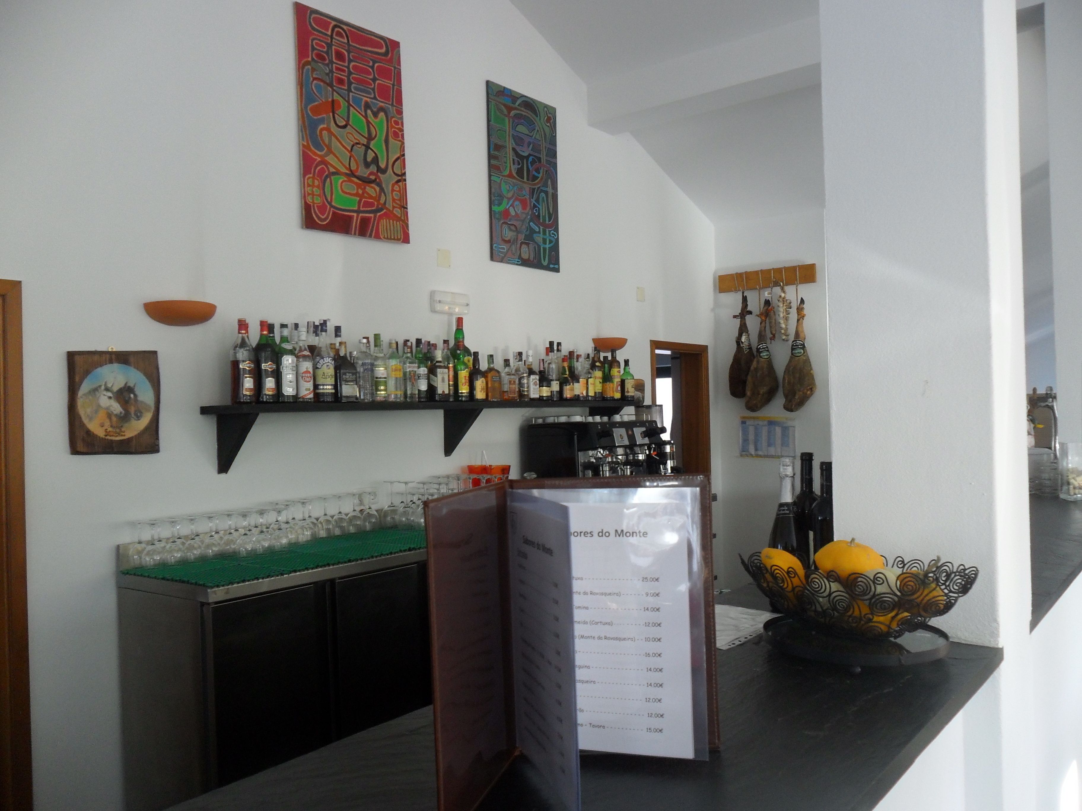 Zona de bar con estantes llenos de botellas de licores, un tablero de menú y elementos decorativos como un bol y embutidos colgantes.