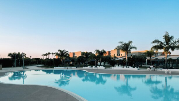 A swimming pool with a curved edge, surrounded by lounge chairs and umbrellas, with buildings and palm trees in the background.