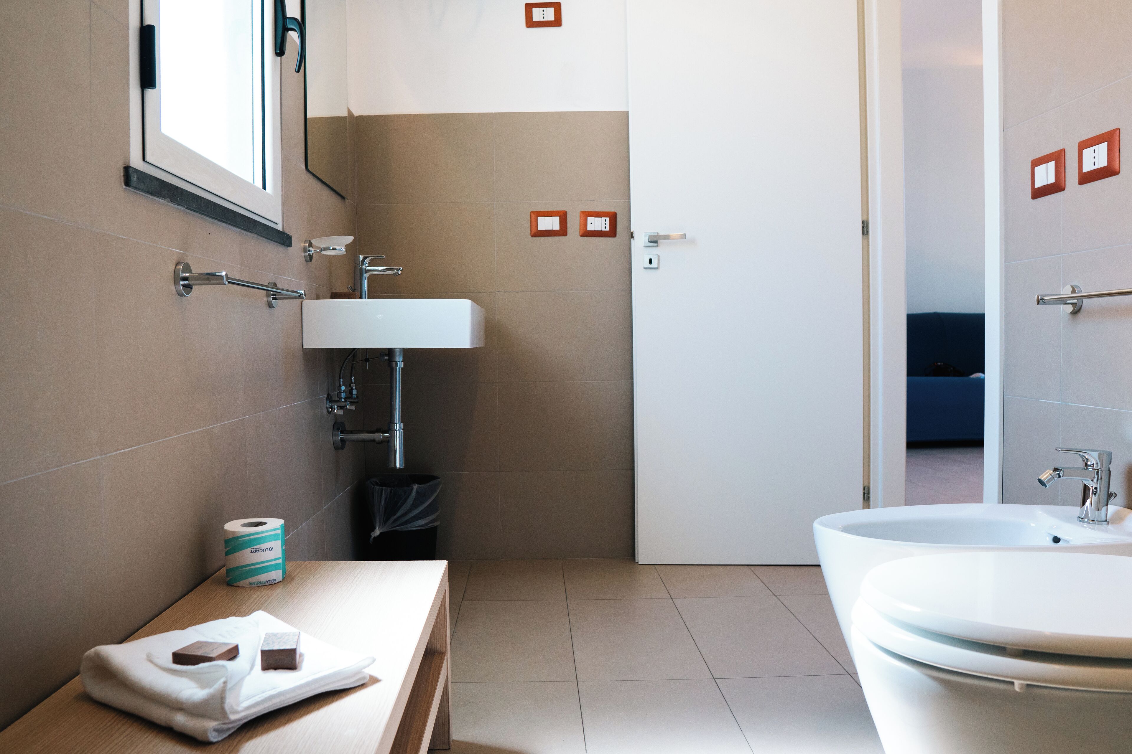 Ein modernes Badezimmer mit Waschbecken, Spiegel und Badewanne. Auf einem Holztisch liegen ein Handtuch und eine Toilettenpapierrolle.