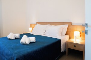 Una cama bien hecha con colcha azul, almohadas blancas y toallas enrolladas al lado. Hay dos mesitas de noche con lámparas.