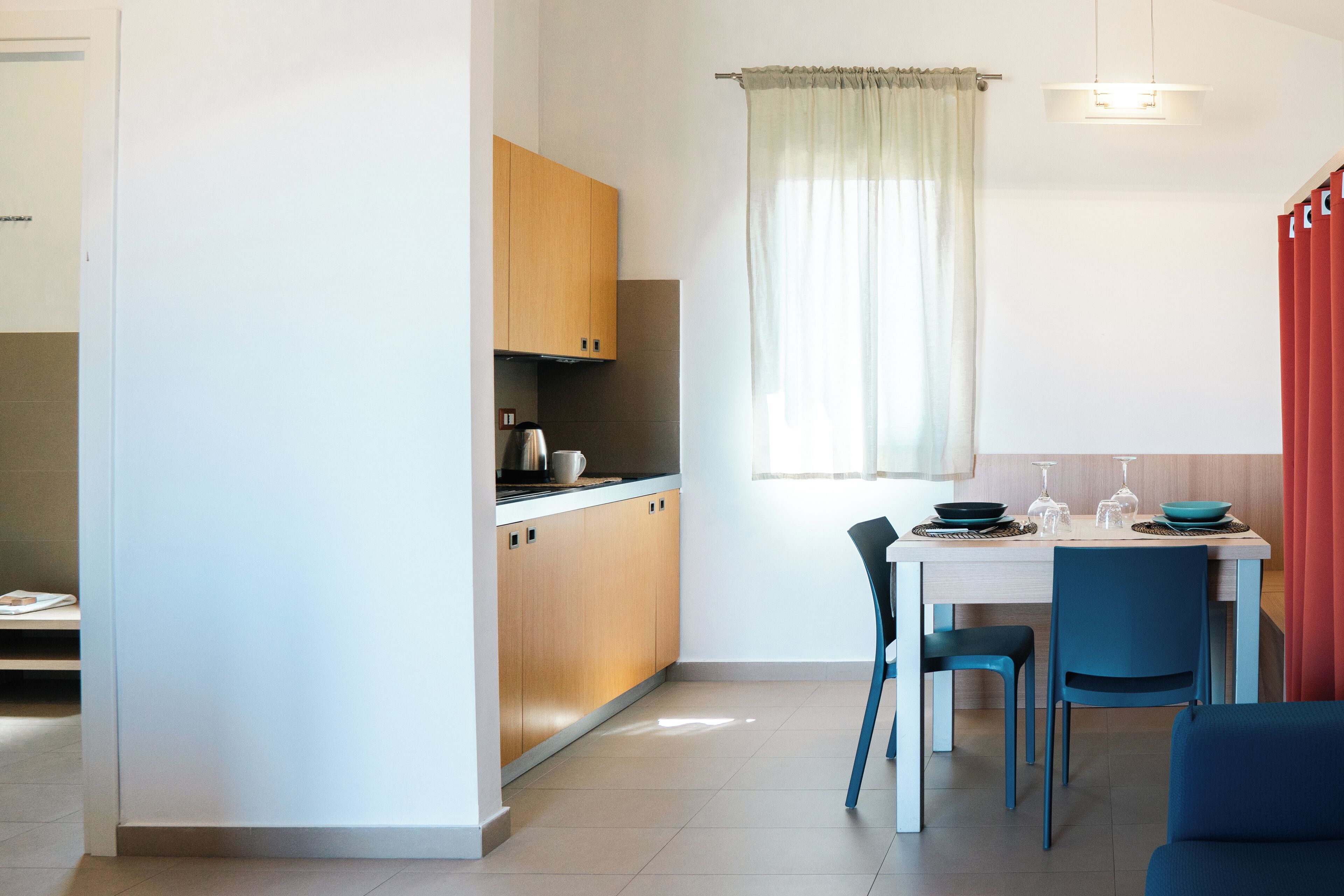 A modern kitchen with wooden cabinets, a dining table with blue chairs, and a window with sheer curtains.