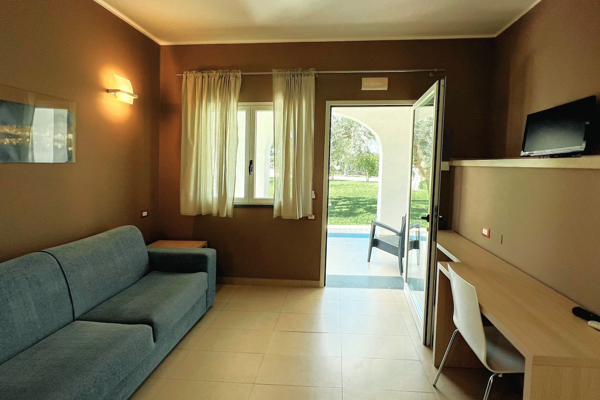 Quarto com sofá, televisão sobre suporte, porta para o exterior e uma pequena mesa.