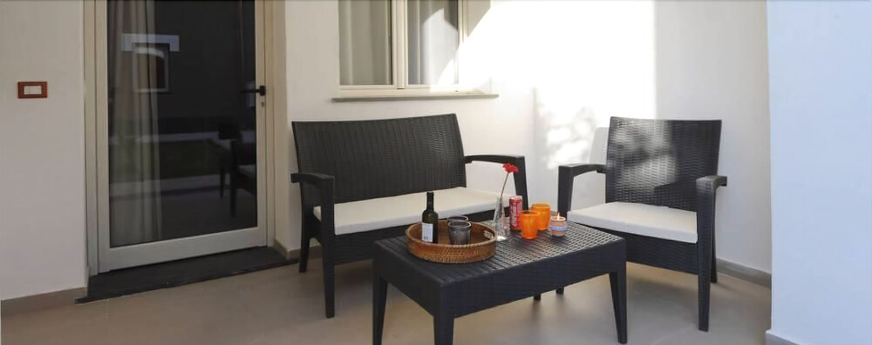 Ein Balkon mit zwei Korbstühlen, einem kleinen Tisch mit Flasche und Kerzen, sowie einer Schiebetür aus Glas.