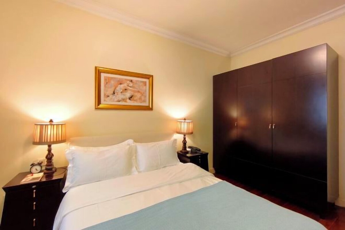 A hotel room with a bed, two bedside lamps, a nightstand, a large wardrobe, and a framed picture on the wall.