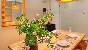 Un comedor con una mesa de madera dispuesta para dos personas, un jarrón con flores rosadas y dos lámparas colgantes.