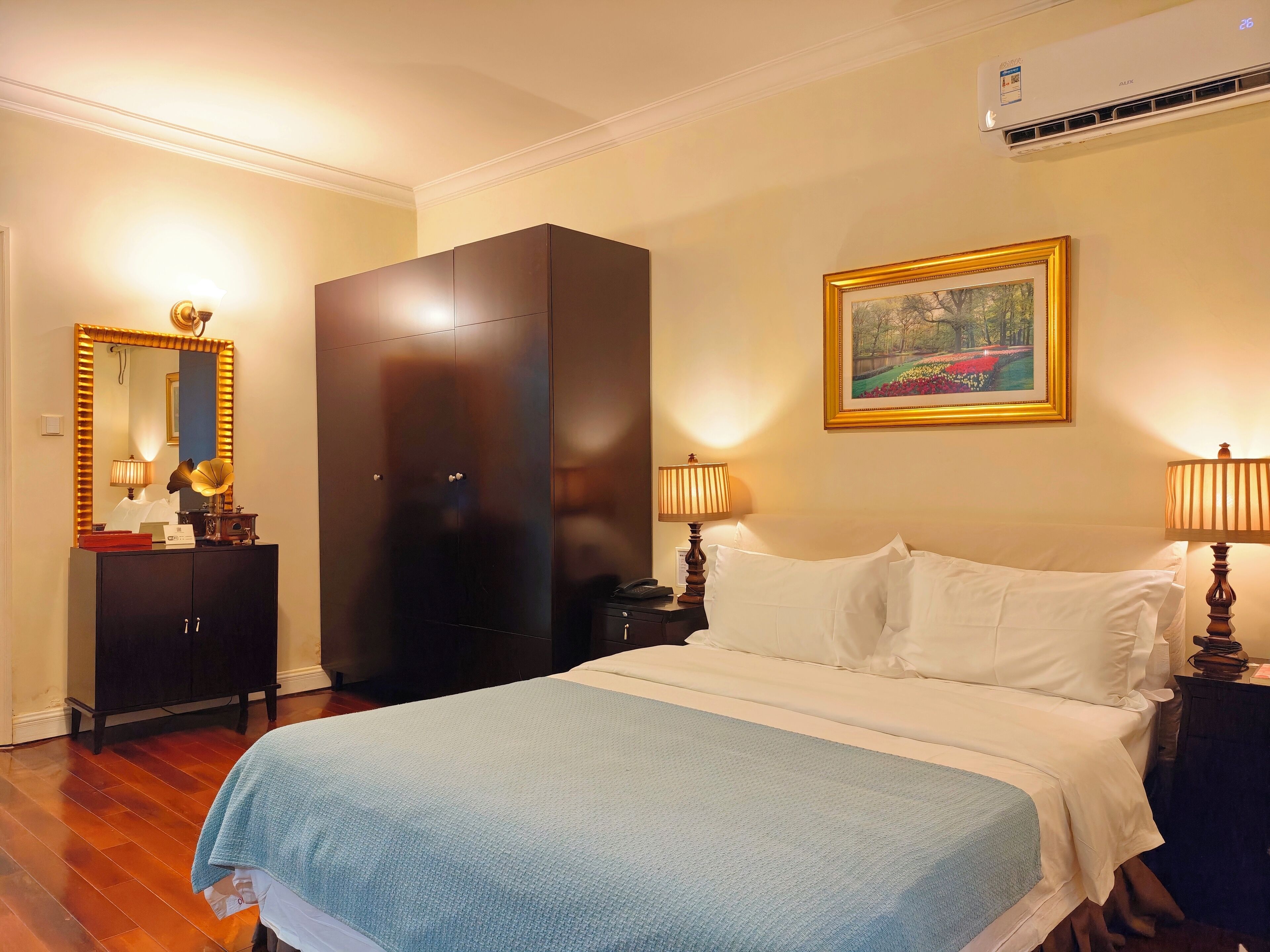 A hotel room with a large bed, a wardrobe, a desk with a lamp, a framed picture on the wall, and an air conditioning unit.