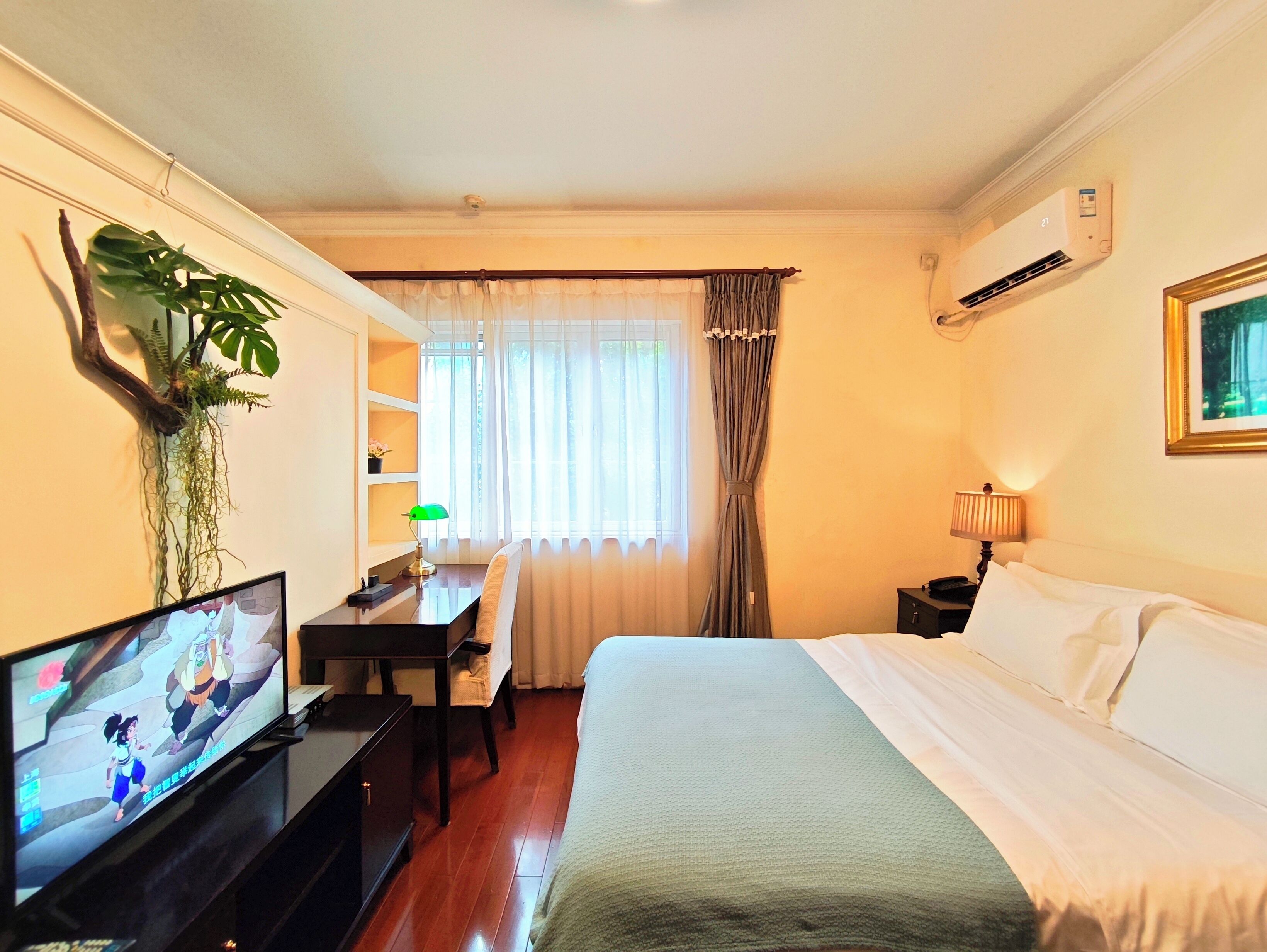 Quarto de hotel com cama, escrivaninha, cadeira, televisão e planta.