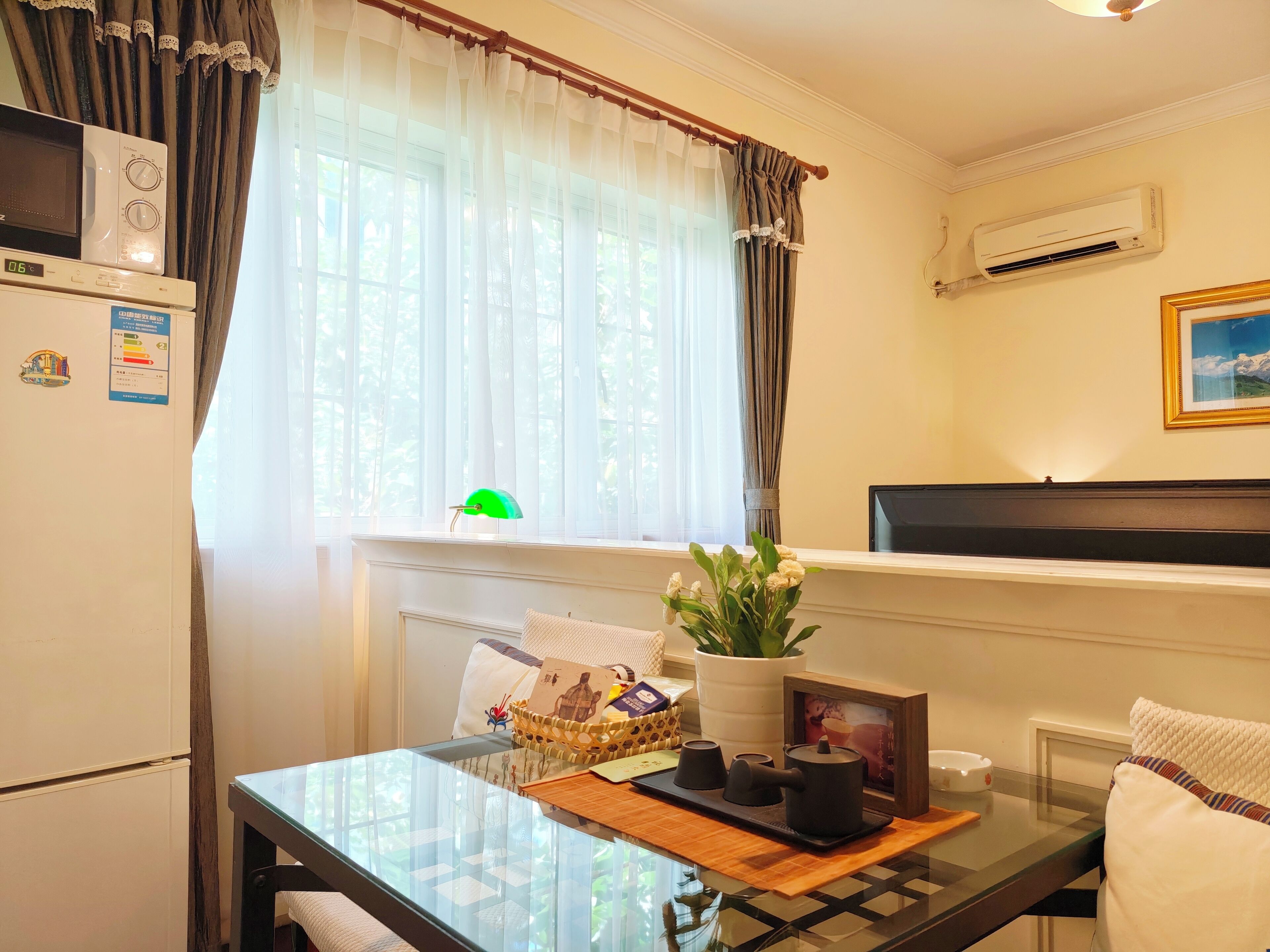 A cozy room with a glass table, a microwave, a refrigerator, a window with sheer curtains, a potted plant, and a framed picture on the wall.