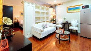 Un salón bien iluminado con un sofá blanco, una mesa redonda de vidrio, un sillón blanco, una mesita auxiliar con una lámpara, una estantería y un refrigerador.