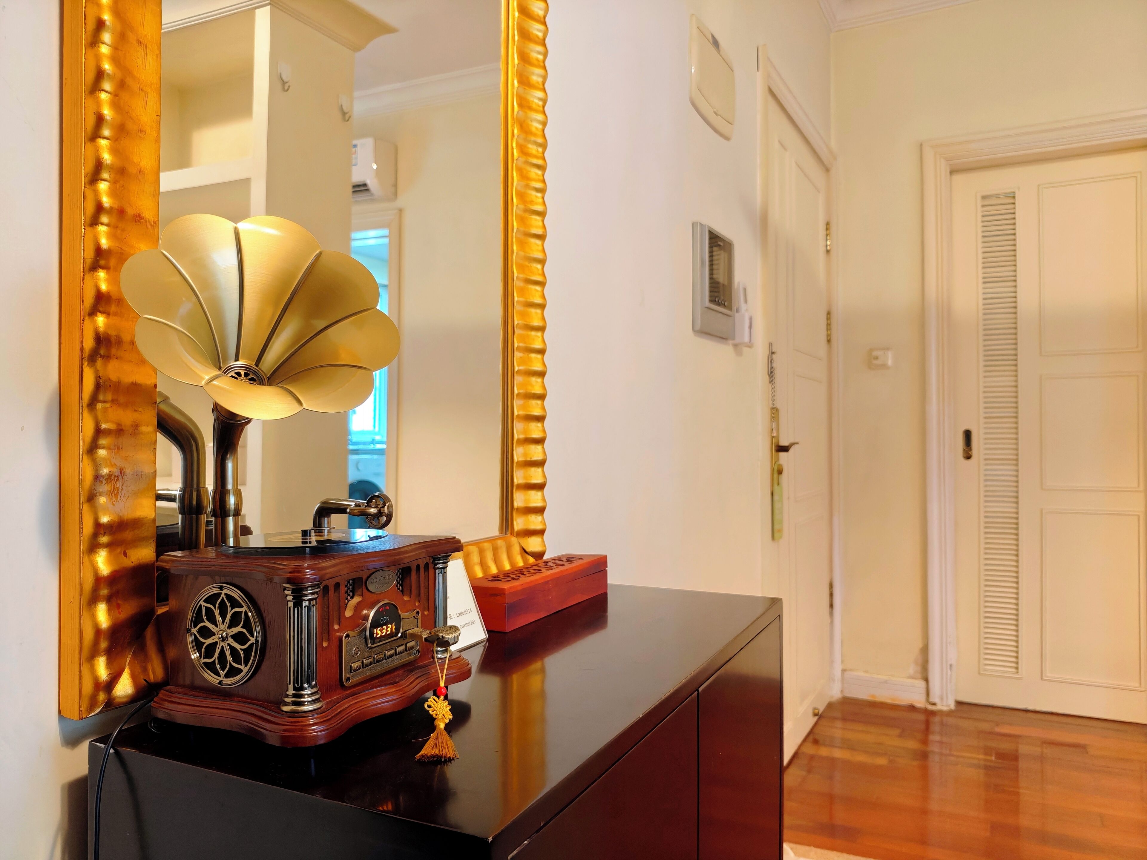 A vintage gramophone on a dark wooden cabinet, a large mirror with a golden frame, and a white door in a hallway.