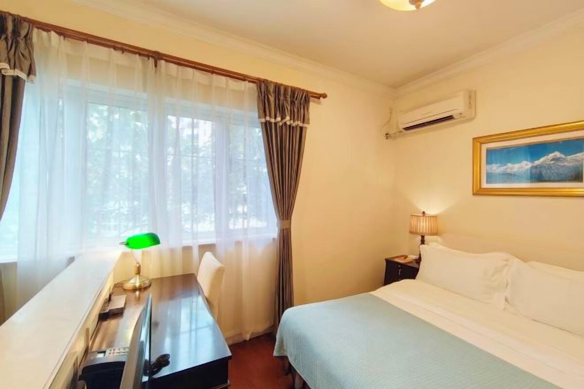 A hotel room with a bed, a desk with a lamp, a chair, and a painting on the wall.