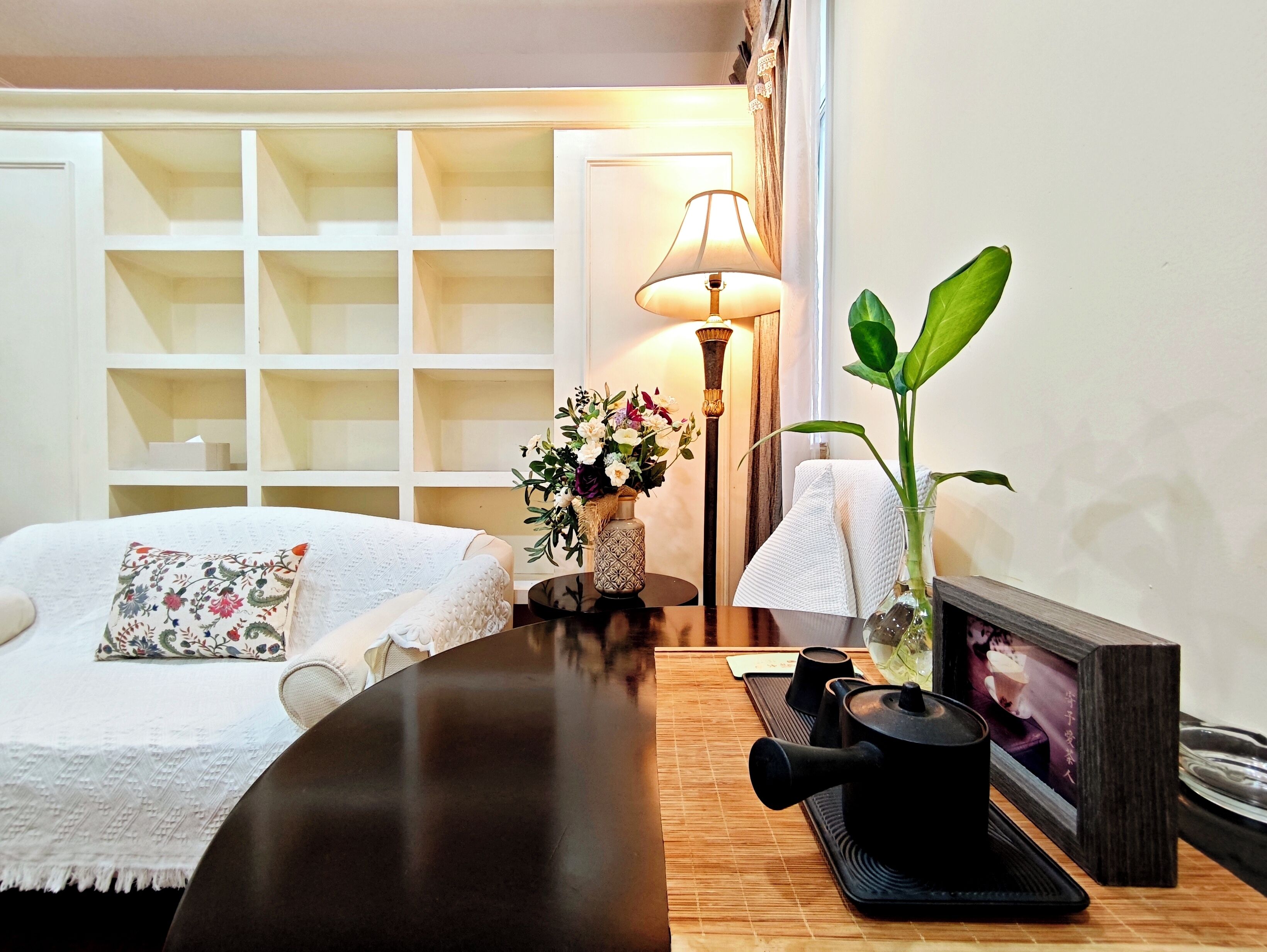 Une chambre d’hôtel bien rangée, avec un lit, une table de chevet sur laquelle se trouve une théière, une lampe et une plante en pot.