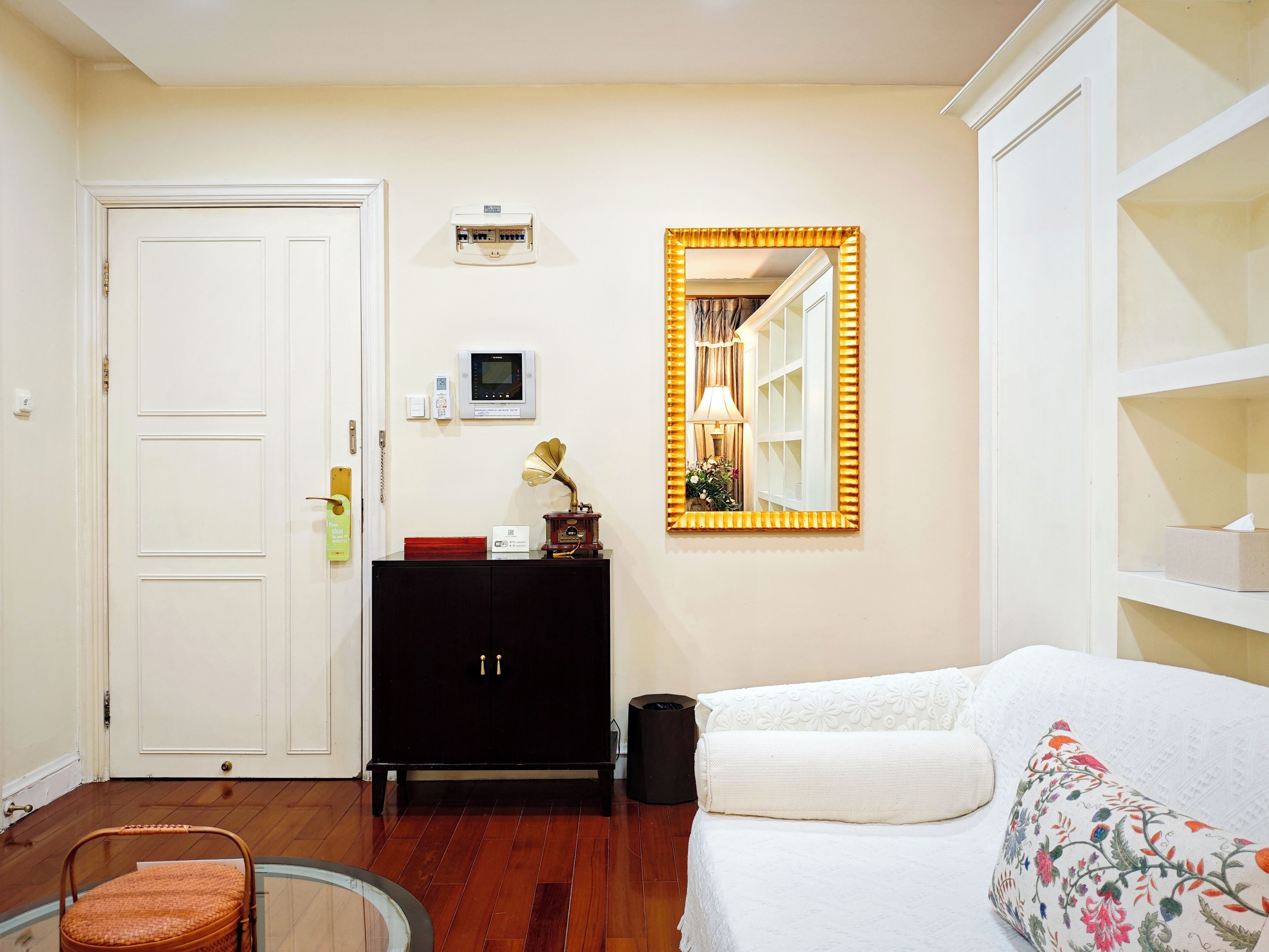 Una habitación con una puerta blanca, un armario negro, un espejo con marco dorado, un sofá blanco y una silla de mimbre.