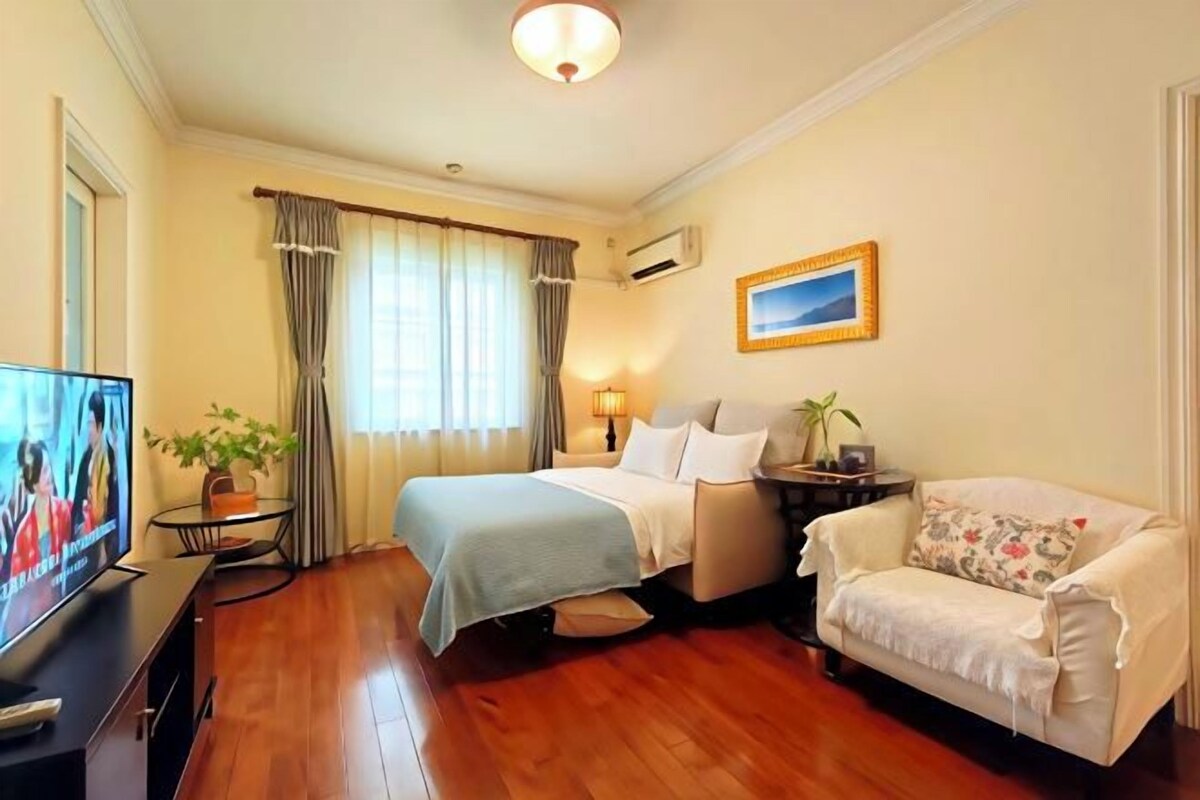 Una habitación de hotel con cama, sofá, televisión, lámpara y ventana con cortinas.