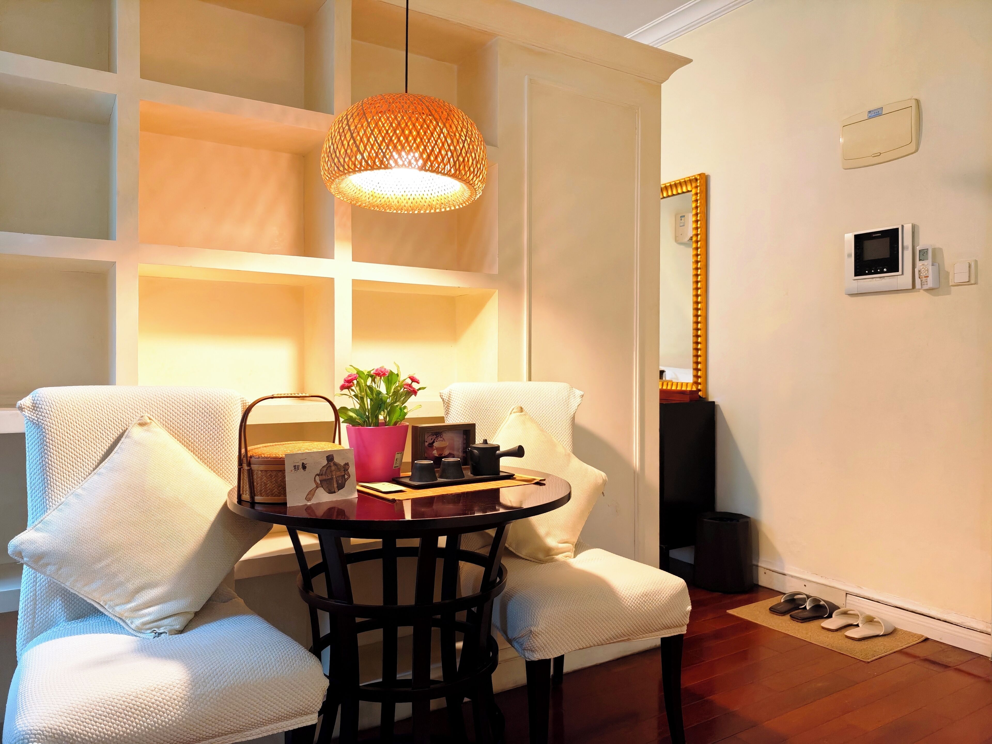 Una habitación con una mesa redonda, dos sillas blancas, una lámpara colgante, un espejo y un termostato en la pared.