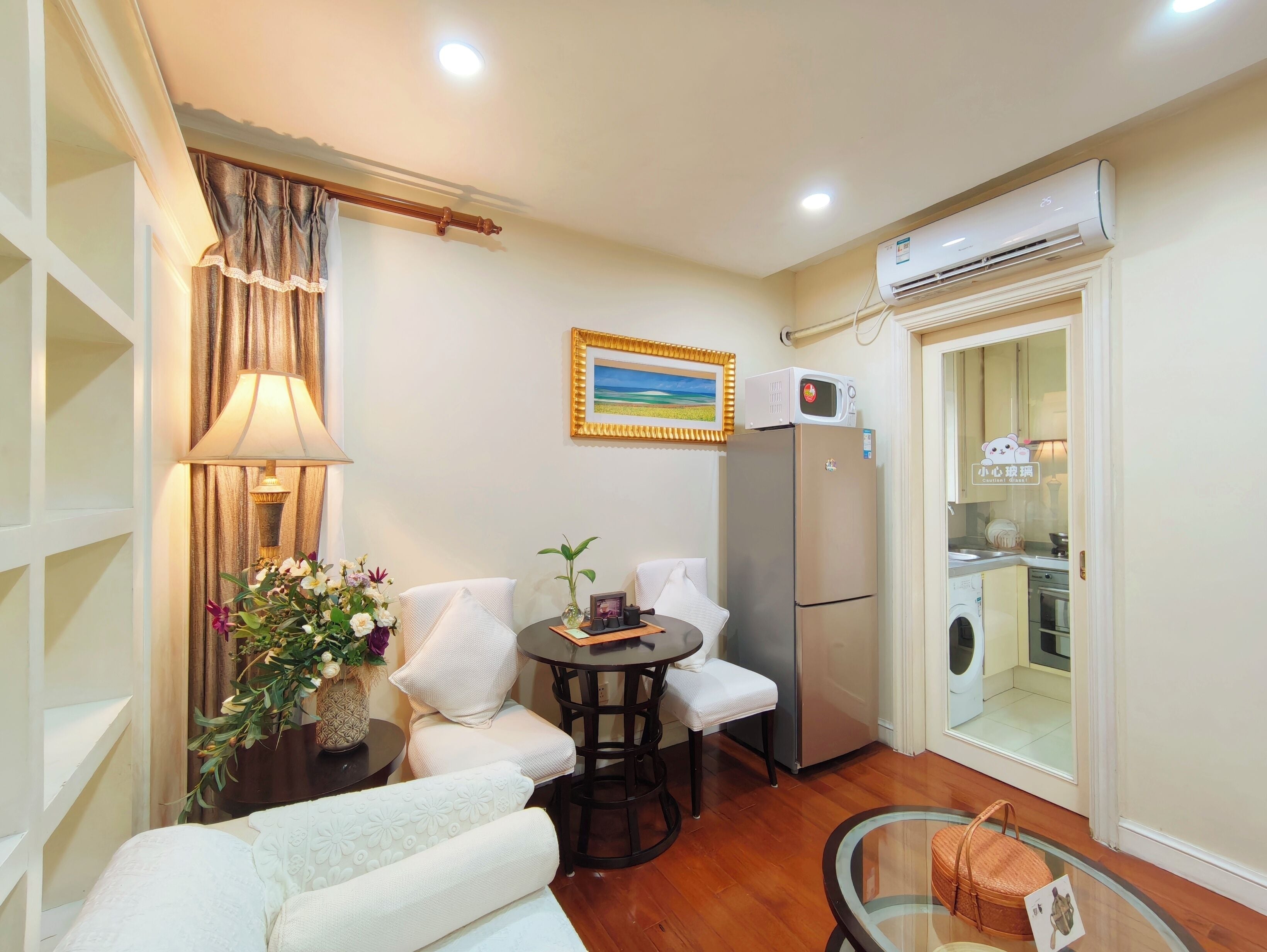 Espacio habitable compacto con sofá, una mesita redonda, una silla, lámpara, planta en maceta, refrigerador, aire acondicionado y lavadora.