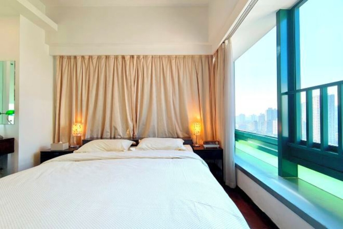 Une chambre d’hôtel avec un grand lit, deux tables de chevet avec des lampes, une vue sur la ville par une grande fenêtre et un rideau qui recouvre la fenêtre.