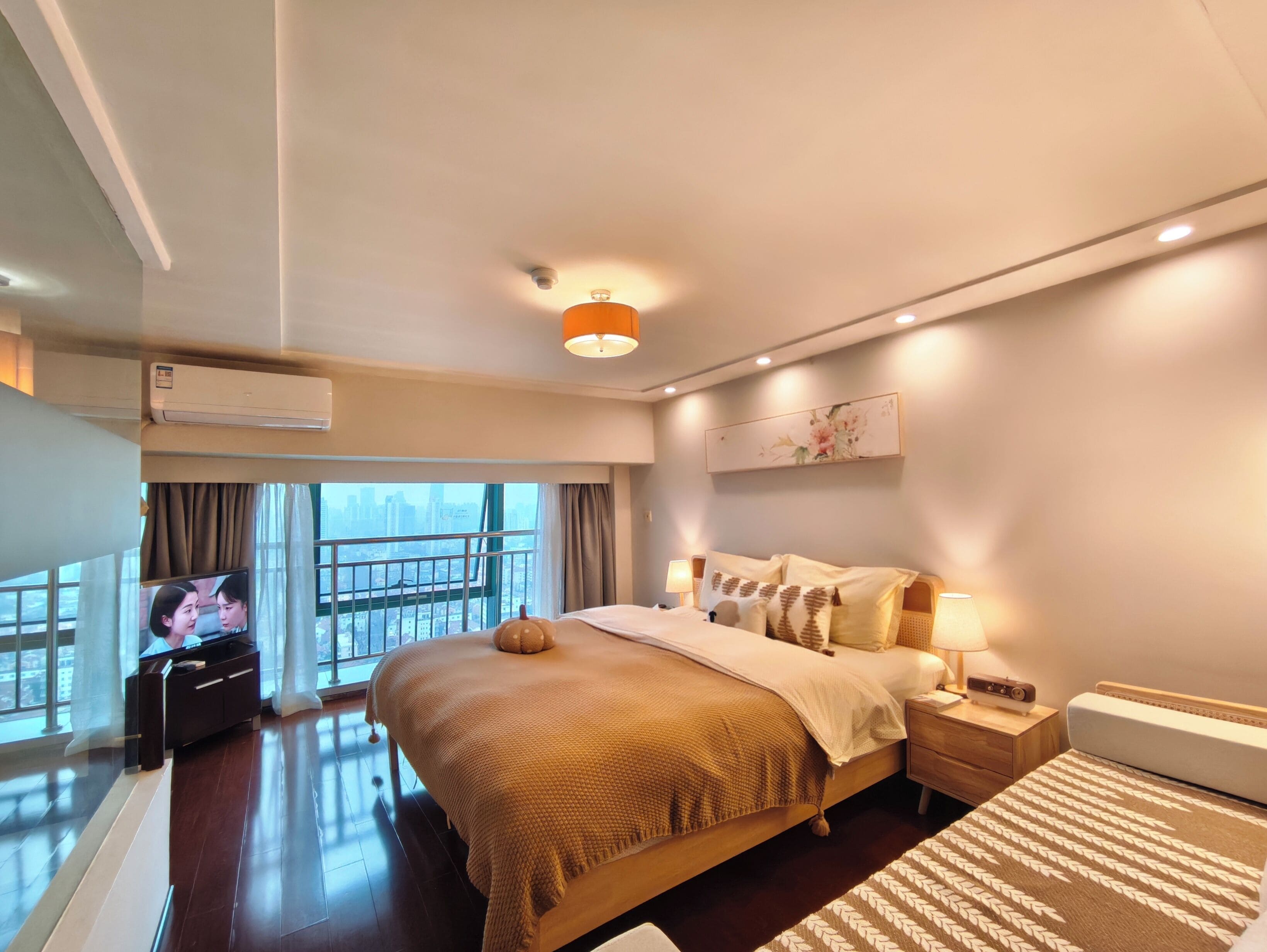 A hotel room with a large bed, a bedside table with a lamp, a TV, and a view of the city through a balcony.