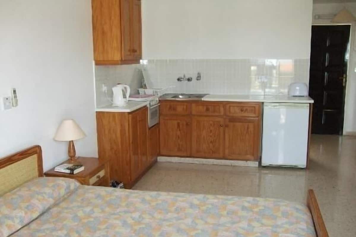 A compact hotel room with a bed, a small bedside table with a lamp, a kitchenette with wooden cabinets, a white refrigerator, and a tiled backsplash.