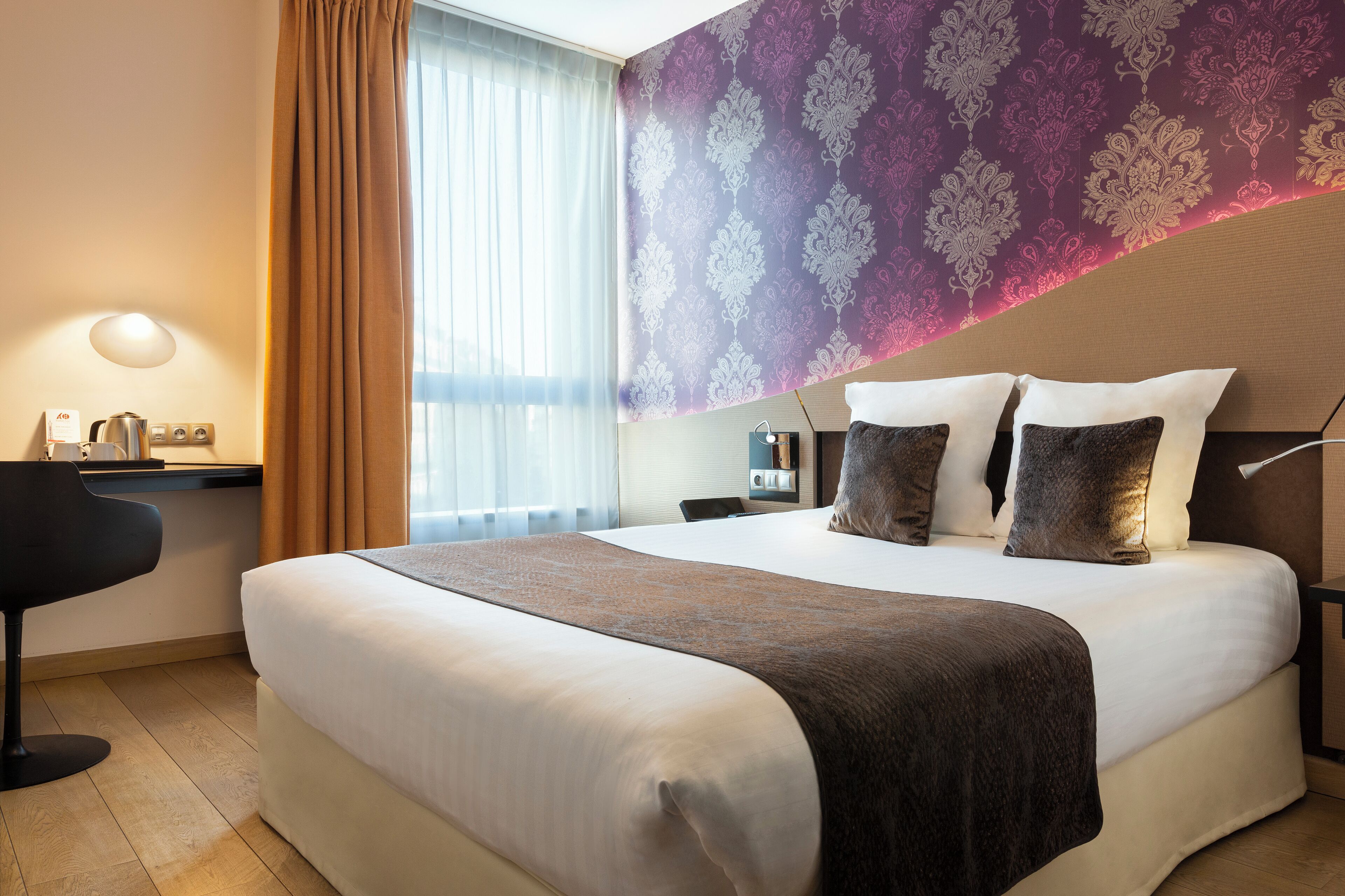 Habitación de hotel con una cama grande, un escritorio con lámpara, una silla y una ventana con cortinas.