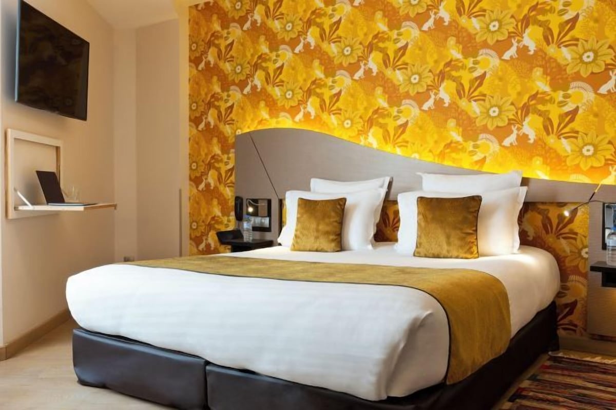 A hotel room with a large bed, a headboard, a bedside table, and a wall with a floral pattern.