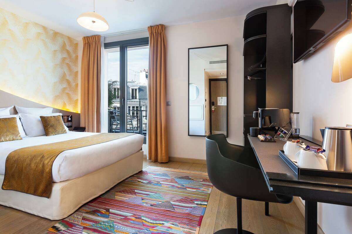 A hotel room with a bed, a desk, a chair, a coffee maker, and a large window with curtains.
