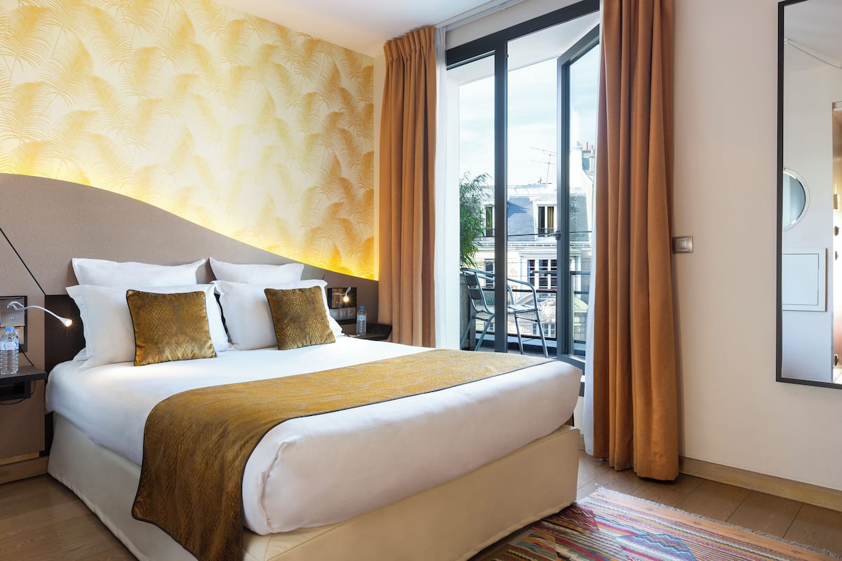 A hotel room with a large bed, a nightstand, a lamp, and a view of the city through a large window.