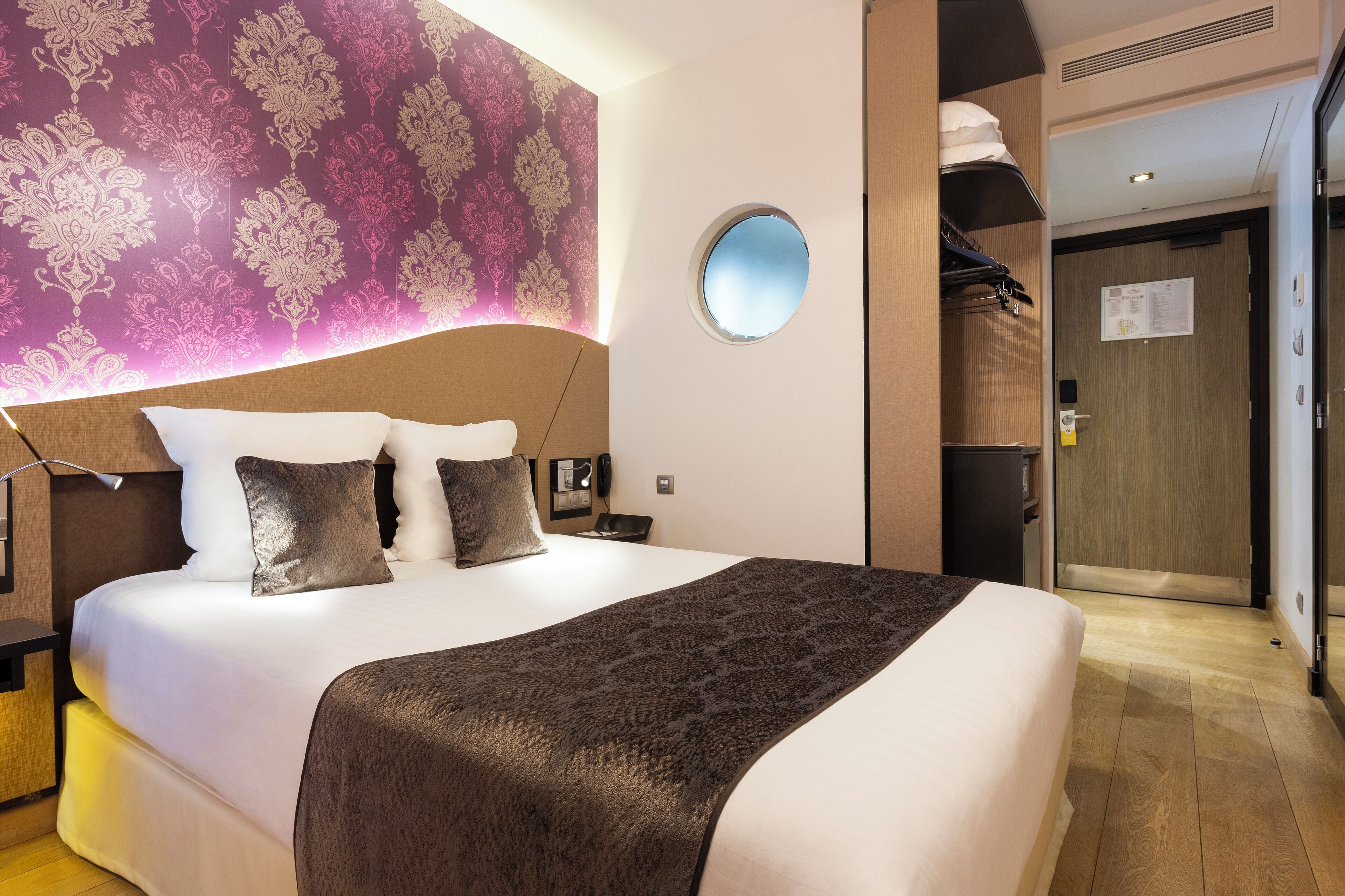 A hotel room with a large bed, a round mirror, a wardrobe, and a patterned wall.