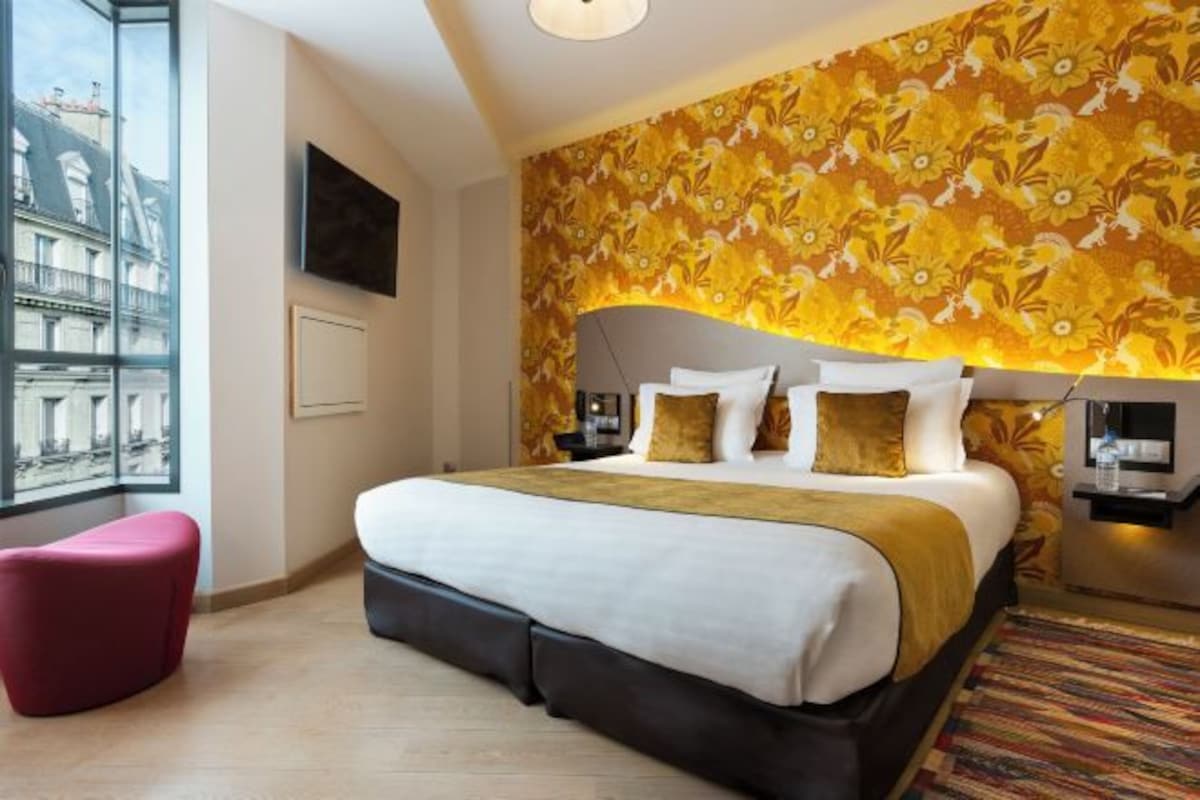 Habitación de hotel con una cama grande, televisor, ventana con vista a edificios y pared con diseño.