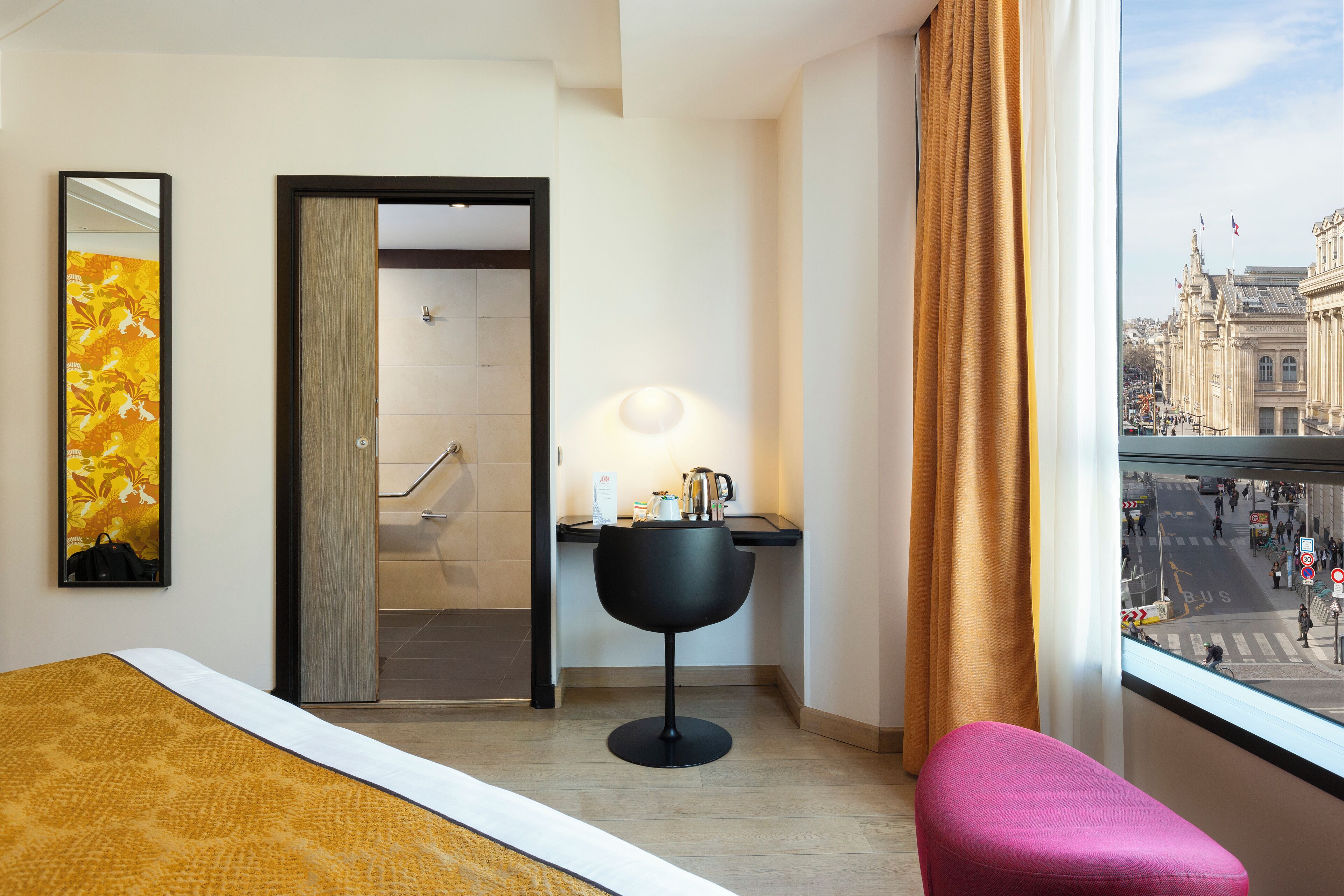 A hotel room with a bed, a desk with a lamp, a chair, a bathroom door, and a view of a city street.