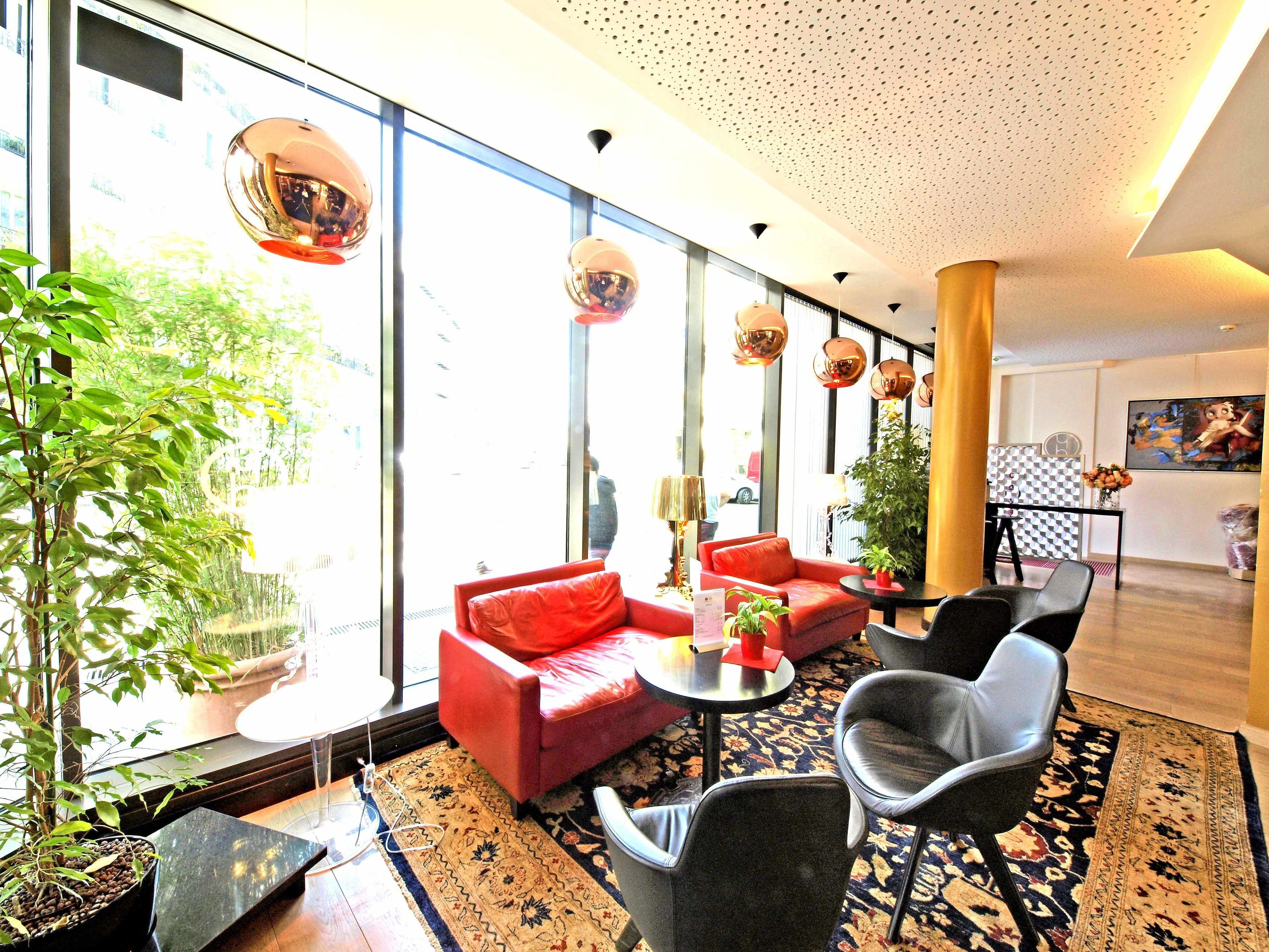 A modern hotel lobby with large windows, red sofas, black chairs, and a patterned rug.