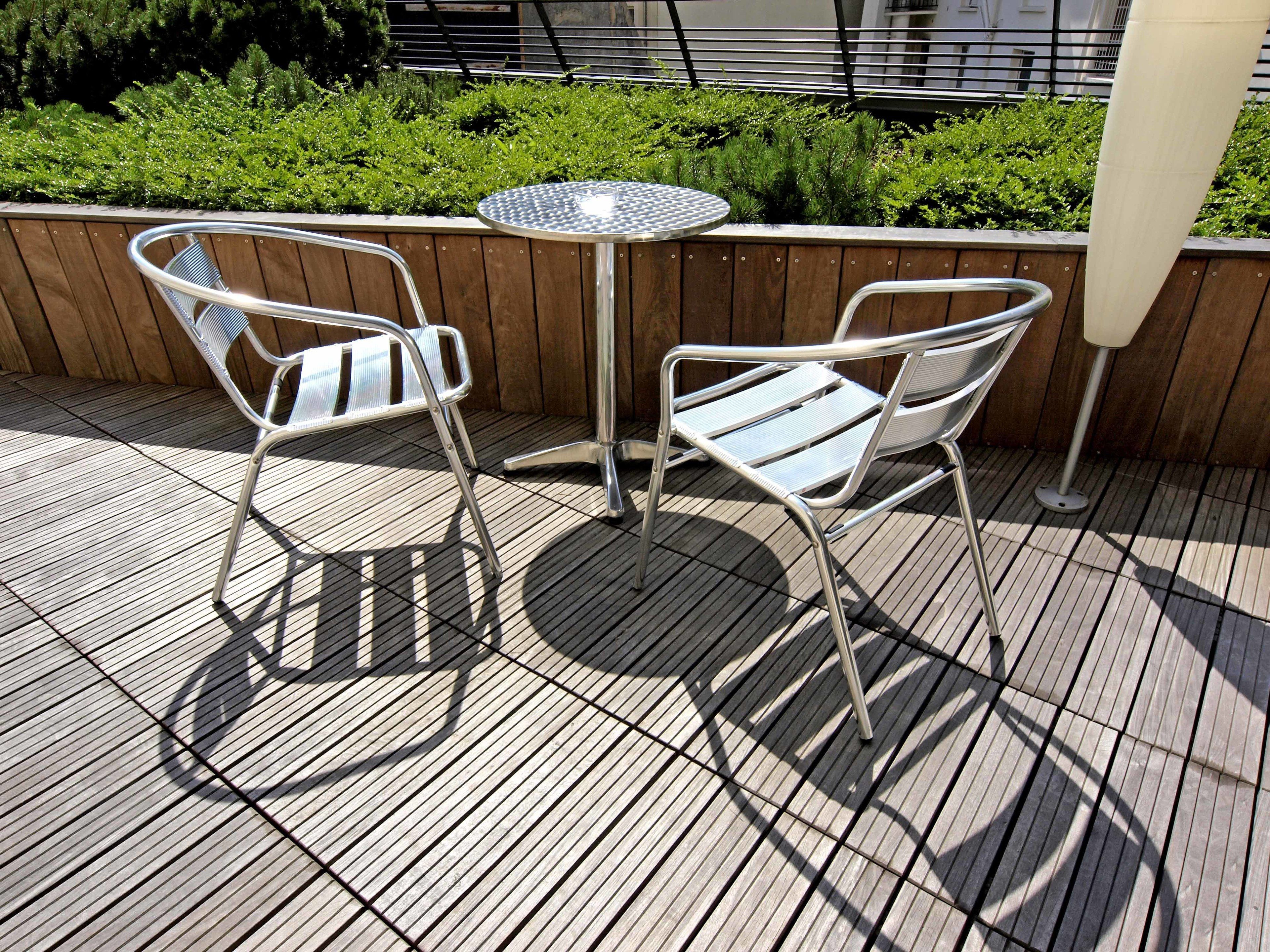 Two metal chairs and a small round table on a wooden deck with a metal railing and greenery in the background.
