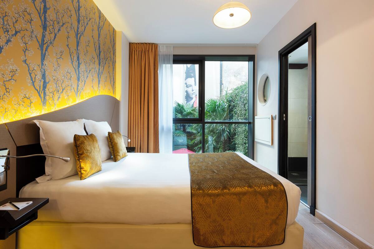 A hotel room with a large bed, a bedside table, a window with a view of greenery, and a door to the outside.