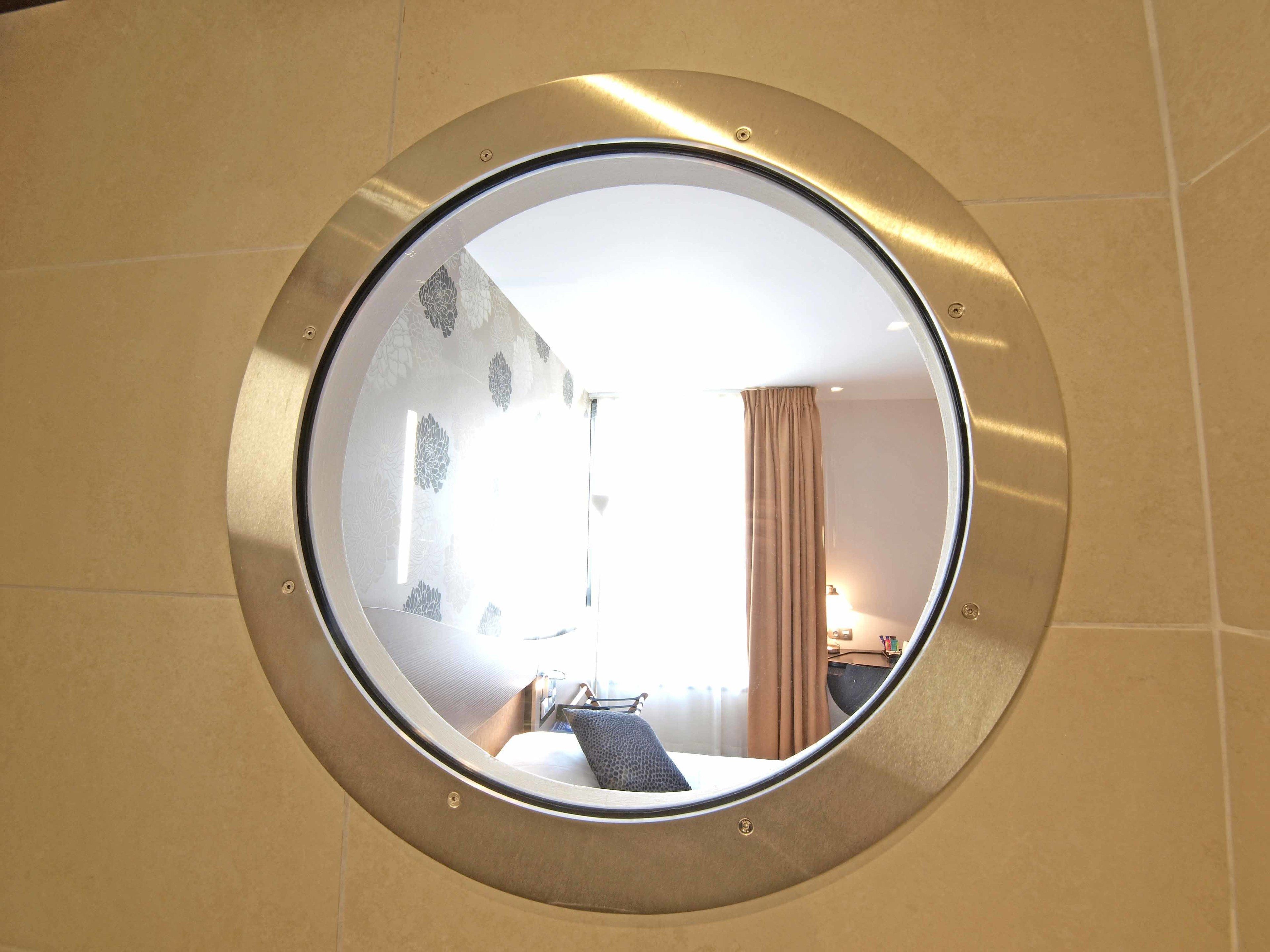 A round mirror with a metallic frame reflecting a room with a bed, a desk, and a window with curtains.
