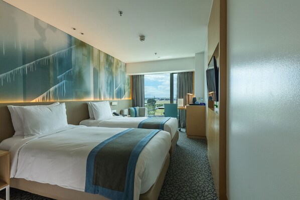 A hotel room with two beds, a large window with a city view, a desk with a chair, and a TV on the wall.