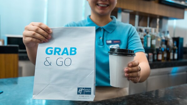 Une personne tenant un sac en papier blanc sur lequel est imprimé le texte « GRAB & GO », ainsi qu’une tasse à café à usage unique.