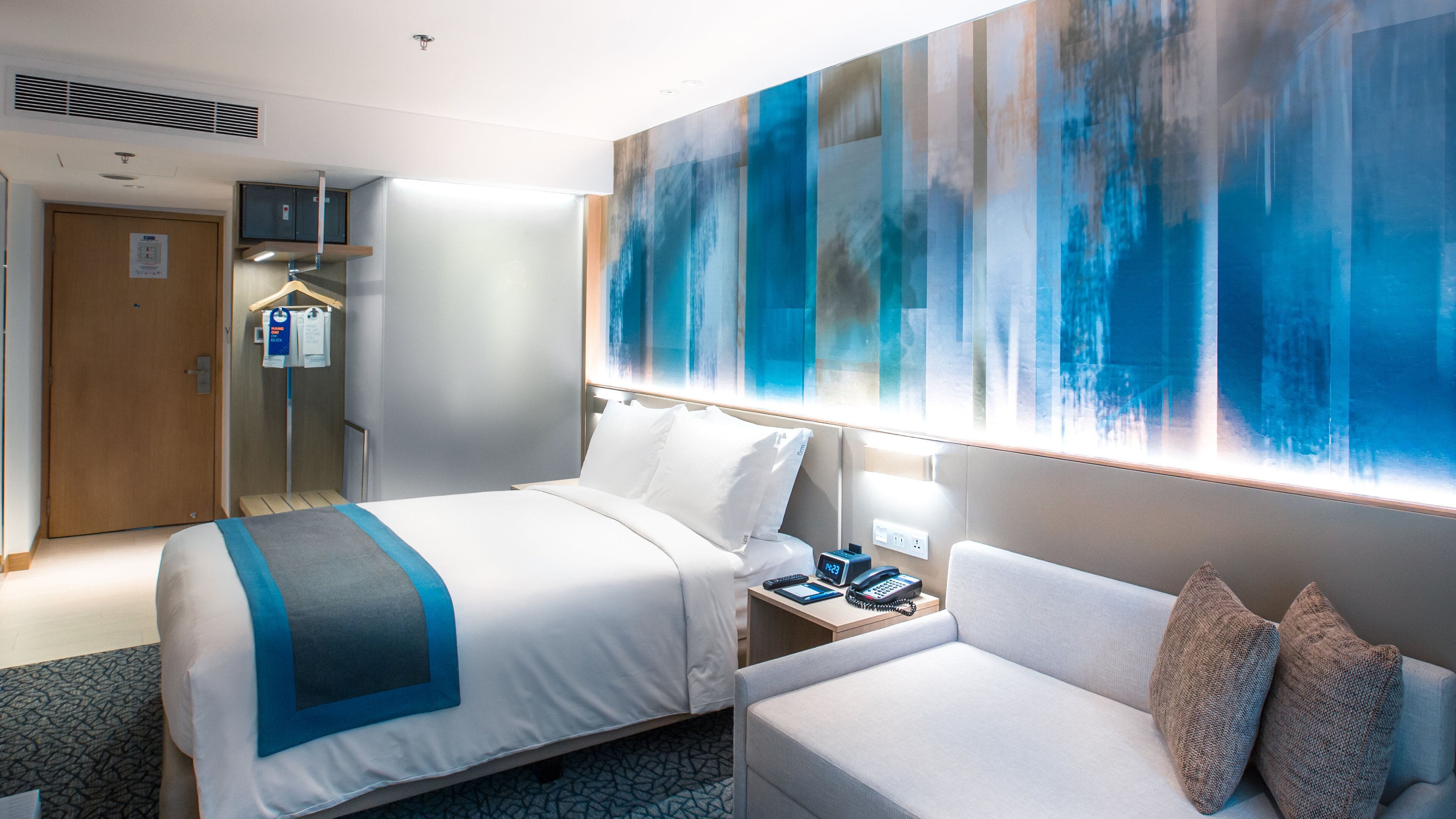 A hotel room with a bed, a sofa, a nightstand, a wall-mounted TV, and a large abstract painting.