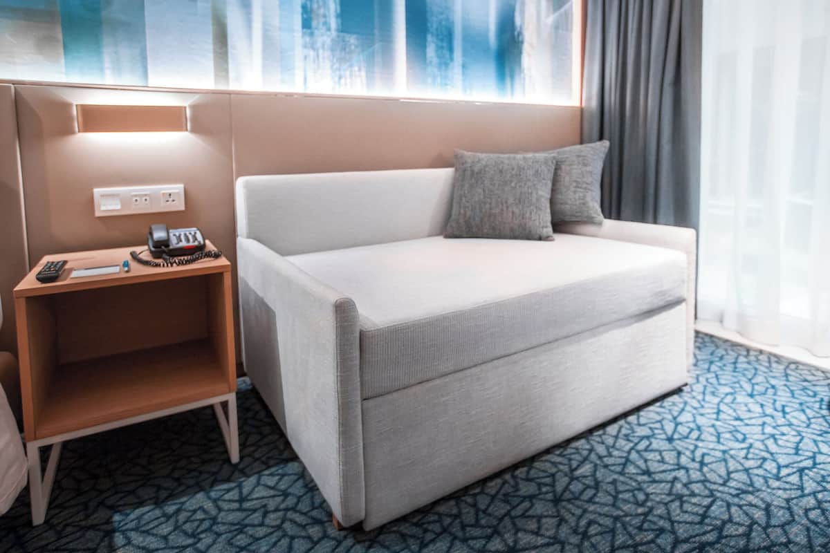 Habitación de hotel con un sofá blanco, una mesita de noche de madera con un teléfono y documentos, y una alfombra estampada.
