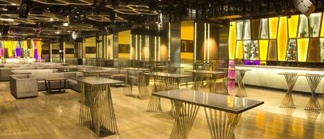 A modern bar area with tables and seating, featuring a unique ceiling design and a bar counter with illuminated shelves.